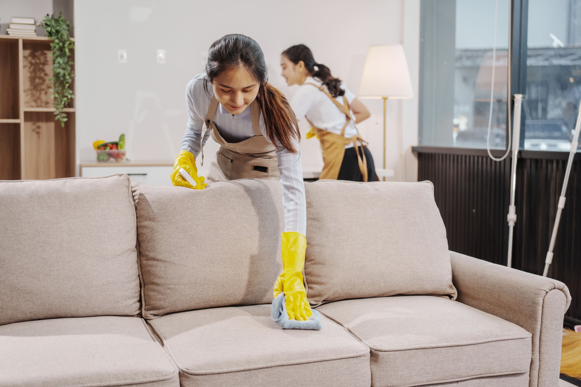 Two women contractors working as housekeepers in an office, wiping glass, sweeping floors, mopping, and cleaning sofas. Providing full-service cleaning for homes, condos, factories, and hotels